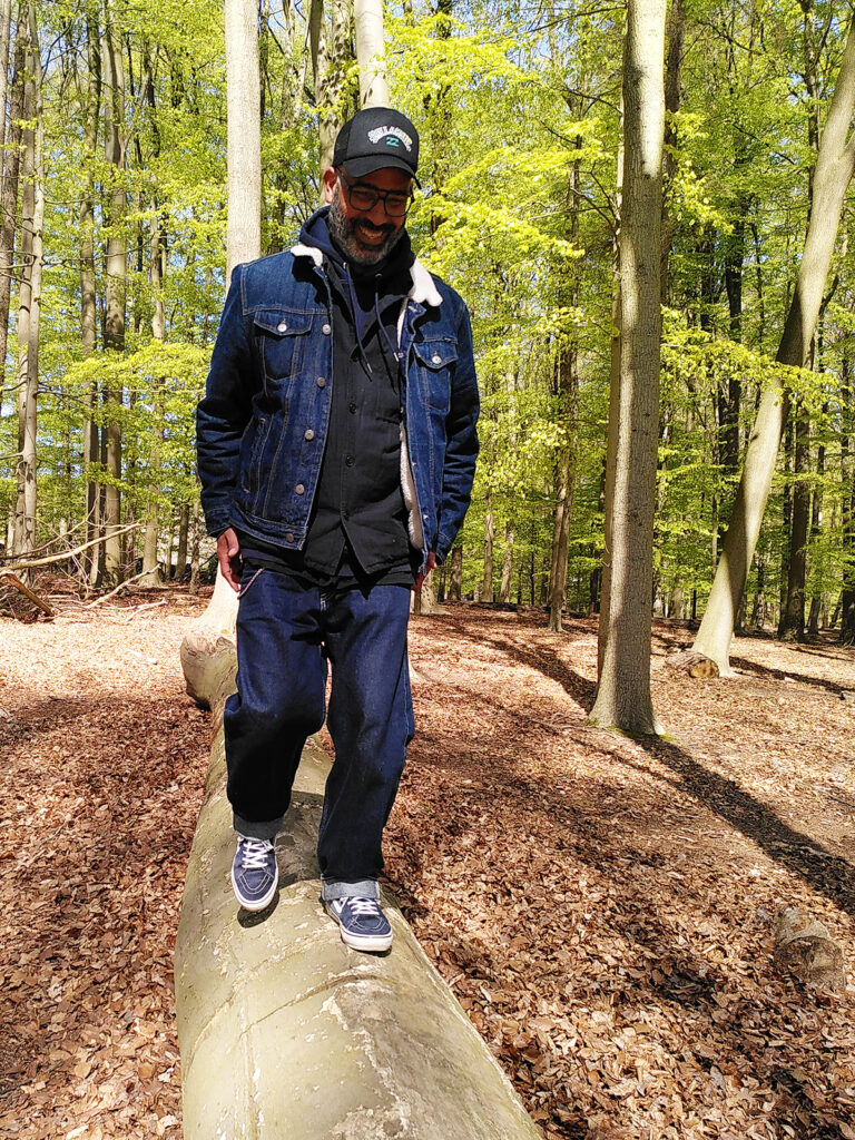 This photo shows Angel Benito Diaz Gonzalez. He is smiling and he is balancing on a tree in the forest and wearing a blue jeans, a denim jacket and a blue Billabong baseball cap.