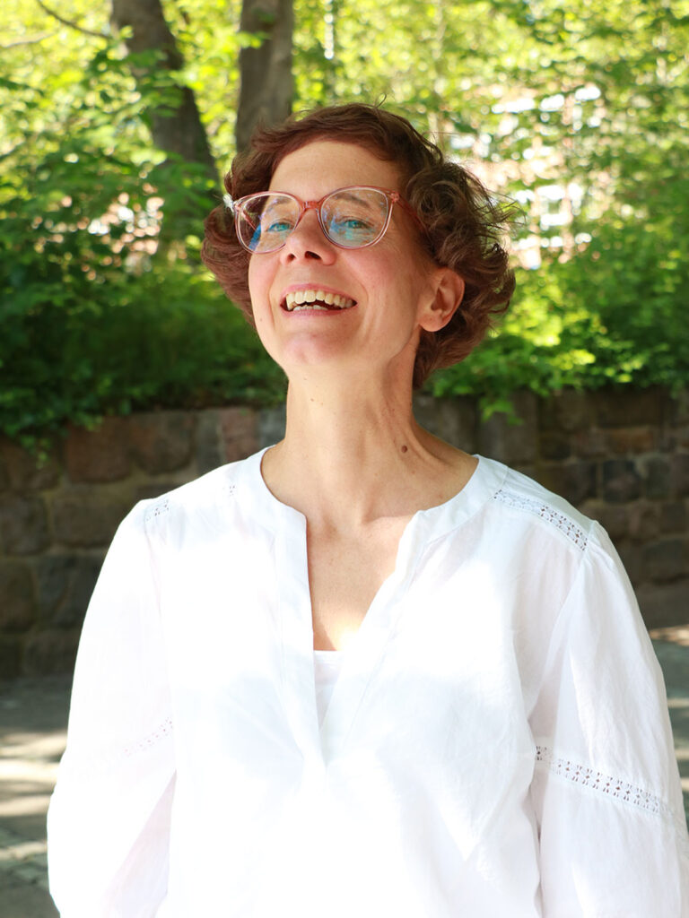 This photo shows Nicole Walter. She has short and curly brown hair, is wearing a white blouse and glasses and she is smiling.