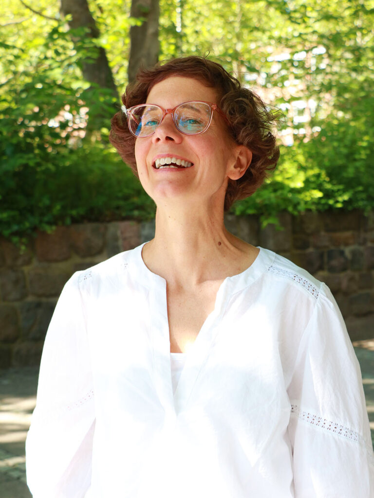 The photo shows Nicole Walter. She is a medium-aged woman with curly, brown hair and glasses. She is wearing a white blouse and in the background there are green trees in the summer sunshine.